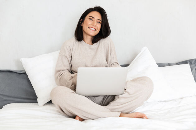 放松一张30多岁的熟女在舒适的公寓里用笔记本电脑 穿着白色亚麻布坐在床上的照片成人笔记本电脑公寓