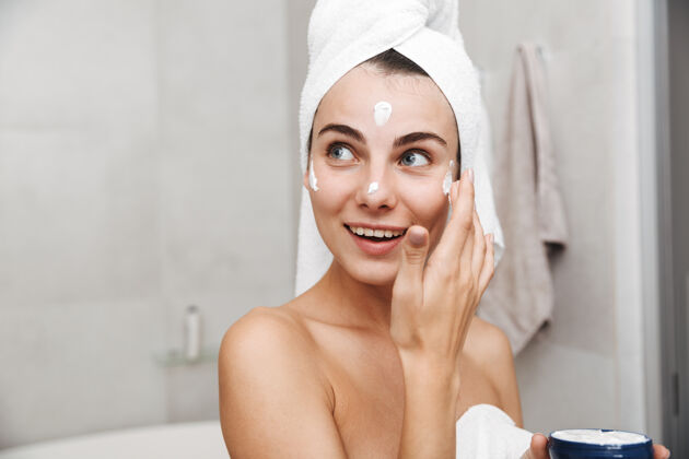 宠爱特写镜头：一个头上戴着毛巾的年轻漂亮女人站在浴室里涂面霜新鲜卫生过程