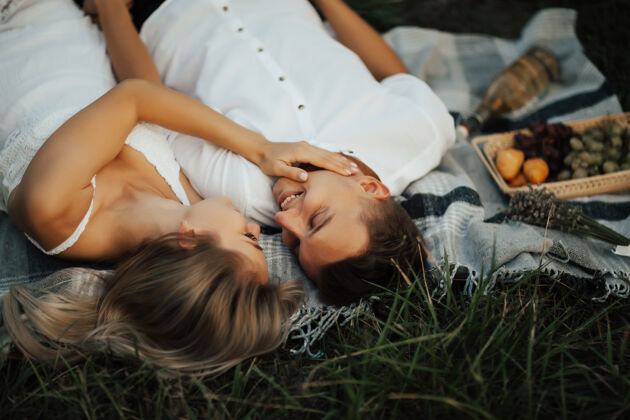 微笑公园里的一对幸福的年轻夫妇正在夏天放松野餐他们躺在绿草上的毯子上 面带微笑地看着对方金发举行季节