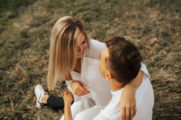户外一对穿着白色衣服的年轻夫妇坐在夏日公园的绿草上 拥抱着 微笑着女孩快乐激情