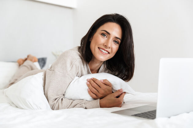 消遣一张30多岁的微笑女人在明亮的房间里躺在白色亚麻布床上使用笔记本电脑的照片可爱舒适满足