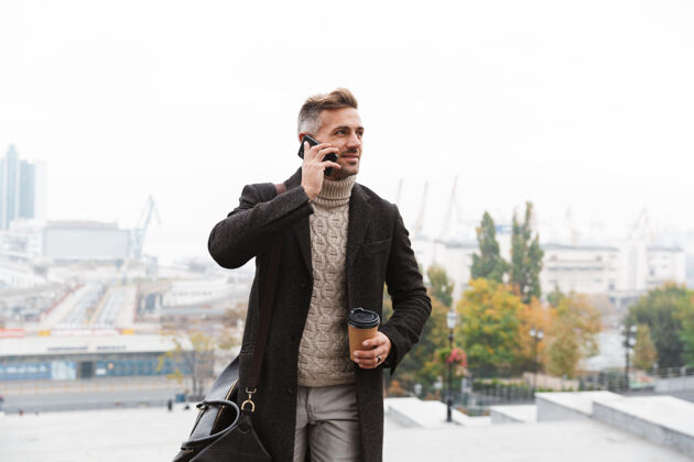 站立30多岁的白种人穿着夹克 手持手机 端着外卖咖啡 走在城市街道上漫步手机户外