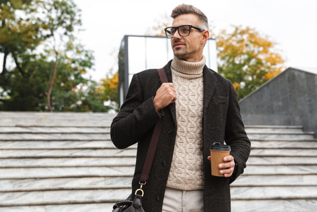 成熟30多岁的微笑男人戴着眼镜 穿过城市街道 拿着外卖咖啡咖啡肖像城镇