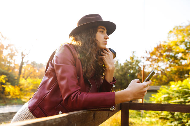 谈话年轻的卷发女人在秋天的公园里用手机喝咖啡手机热秋天