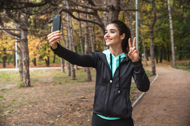 公园在公园里 一位年轻漂亮的健身女士在户外用手机拍下了一张充满和平姿态的照片白种人花园采取