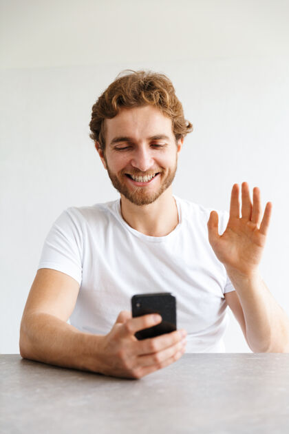 成人照片中 一个快乐的留着胡子的年轻人在家里的桌子旁用手机挥手交谈短信手机年轻