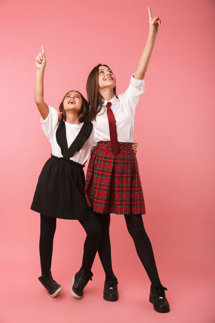 小学生两个穿着校服的开朗女生孤立地站在粉红色的墙上 手指着友谊时尚站着