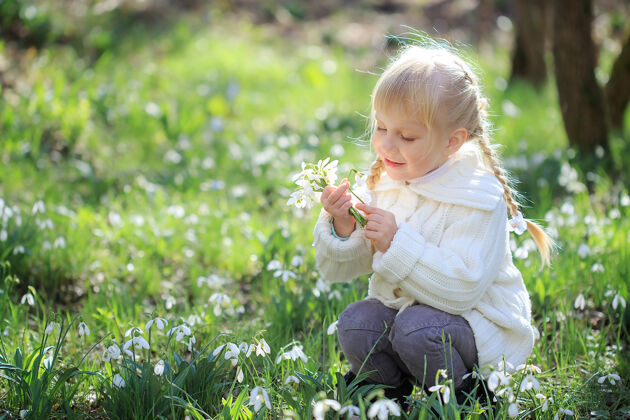 公园一个漂亮的小女孩正坐在一片鲜花盛开的草地上一个穿着白色针织毛衣的小女孩正在考虑放雪球复活节时间春天阳光明媚的森林白天草地欢呼