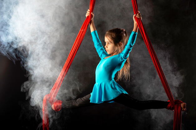 运动一个穿着蓝色运动服的漂亮小女孩在空中做了一个裂口 她紧紧抓住一条被黑烟包围的红色飘带年轻吊带飞行