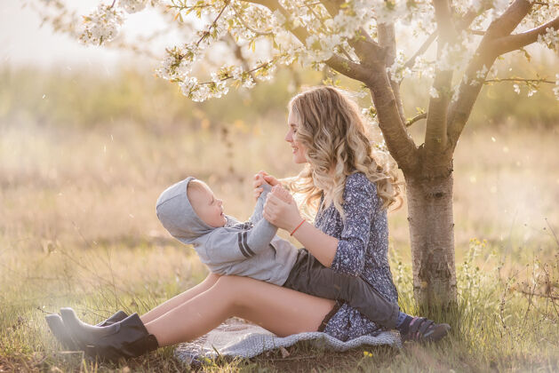 关系年轻的卷发妈妈抱着儿子 坐在绿草旁盛开的白樱桃树上开花阳光男孩