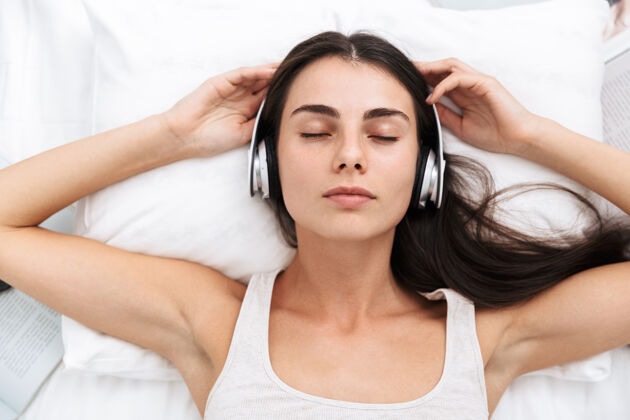 视图俯视图：一位美丽的年轻女子躺在床上用耳机听音乐音乐清醒电子