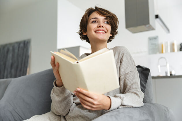 室内快乐的年轻女子坐在家里的沙发上看书年轻聪明休闲
