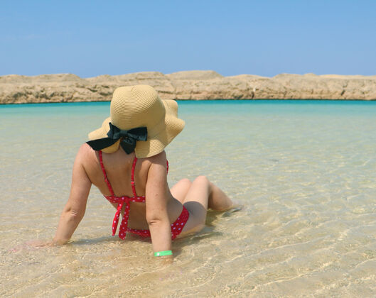 泳衣一个穿着泳衣 戴着帽子的女孩坐在海水中 背景是岩石帽子夏天天空