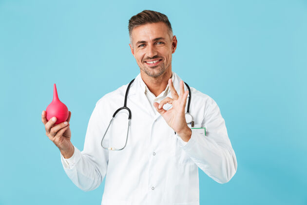 帅哥快乐的医学专家穿着白大褂拿着灌肠器 孤立地站在蓝色的墙上心脏病专家制服医生
