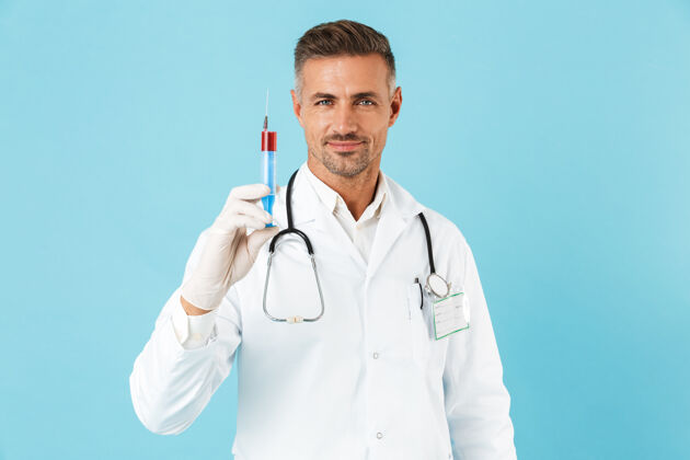 英俊戴着听诊器戴着手套拿着注射器的快乐医生的画像 孤立地站在蓝色的墙上成人男人诊所