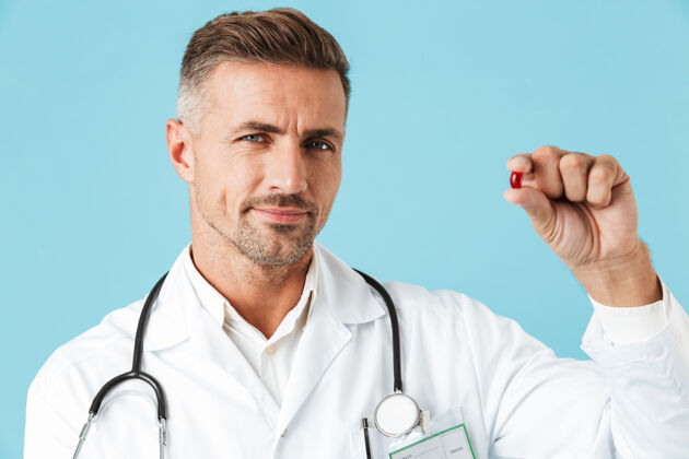 医疗一位身穿白大褂 手拿一颗药丸 孤零零地站在蓝色墙壁上的迷人医生的照片白种人治疗制服