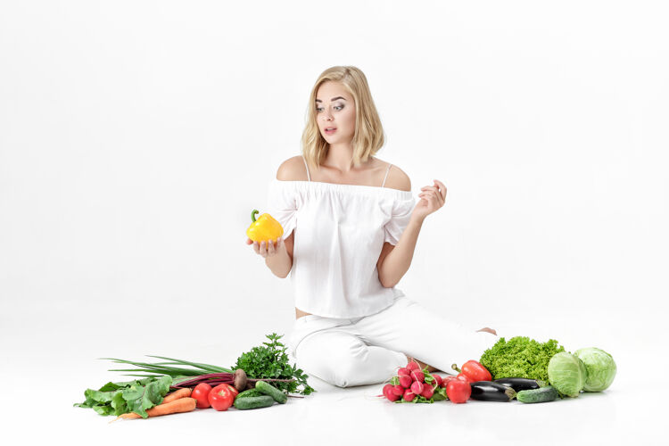 人漂亮的金发女人 穿着白衣服 白色的桌子上放着很多新鲜蔬菜背景女孩拿着甜椒消费者有机背景