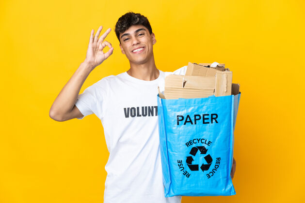 乐观一名男子拿着一个装满废纸的回收袋 在隔离的黄色墙壁上用手指画着“ok”标志 准备回收废纸年轻保护减肥