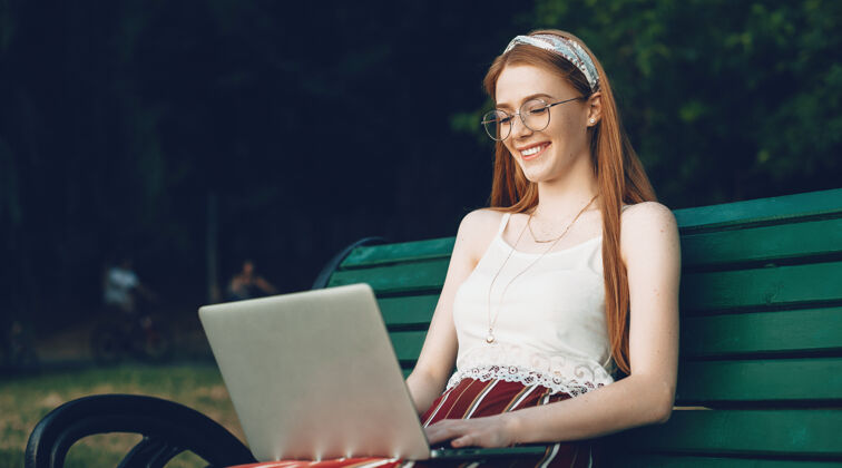 雀斑一位长着雀斑的姜黄色高加索女士正在公园的长凳上对着笔记本电脑打字 她笑了使用生姜红发