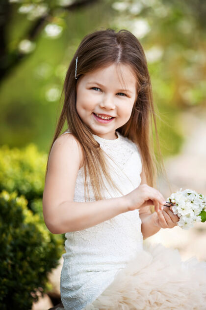 小美丽微笑的小女孩穿着奶油色的裙子 映衬着夏日公园的绿色年轻花春天