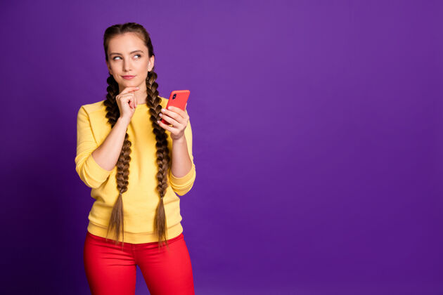 女性令人惊叹的少女手持电话寻找空旷的空间有创意的新想法后穿休闲黄色套头衫红色裤子隔离紫色墙壁浏览长电话