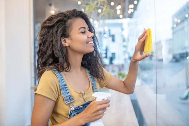 清洁咖啡馆 清洁年轻在咖啡馆用黄色餐巾纸擦窗户的轮廓里 长着波浪形头发的漂亮女人忙碌工作日关注