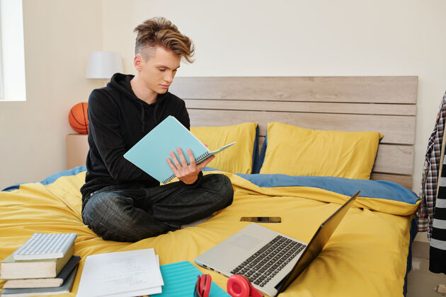 欢呼软件工程专业的学生坐在床上 拿着书 笔记本电脑和课本 在笔记本上写字学生学习学习