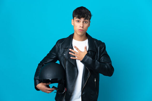 拉美裔一个戴着摩托车头盔的年轻人在隔离的蓝色墙壁上看着右边 惊讶和震惊皮革瘦身驾驶