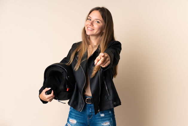 表情立陶宛年轻女子手持摩托车头盔 孤立地站在米色背景上 幸福地指着前方微笑女性司机