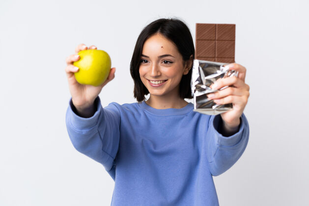 巧克力年轻的白人妇女被隔离在白墙上 一只手拿着巧克力片 另一只手拿着苹果好可可年轻