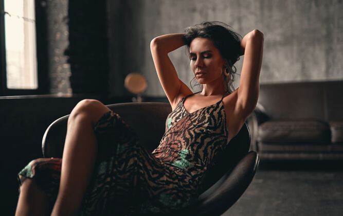 模特一个漂亮的黑发女人的肖像 穿着一件裙子 坐在扶手椅上摆姿势 用手拉直她的长卷发深色再融资姿势