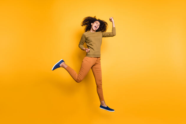 胜利照片紧急卷曲波浪式时尚开朗美丽女朋友穿着橙色裤子鞋子绿色毛衣跳跃奔跑在生动的色彩背景下赢家风扇民族