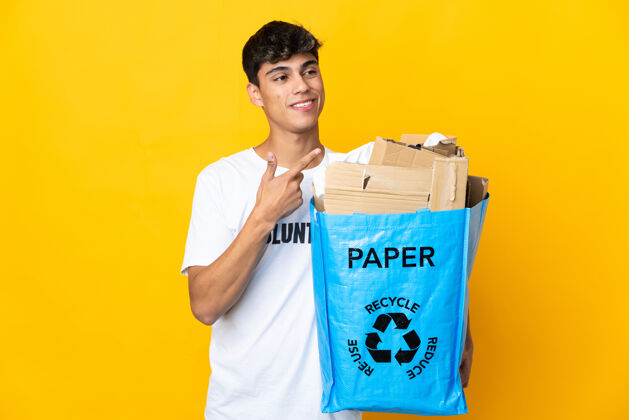 减肥一名男子拿着一个装满废纸的回收袋 翻过隔离的黄色墙壁 指着侧面展示一个产品指点人保护