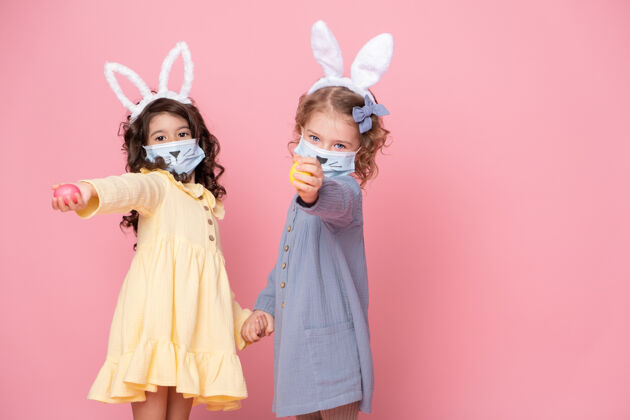 童年两个戴着兔耳头巾和粉红色彩蛋保护面具的女孩背景.covid复活节概念庆祝油漆兔子