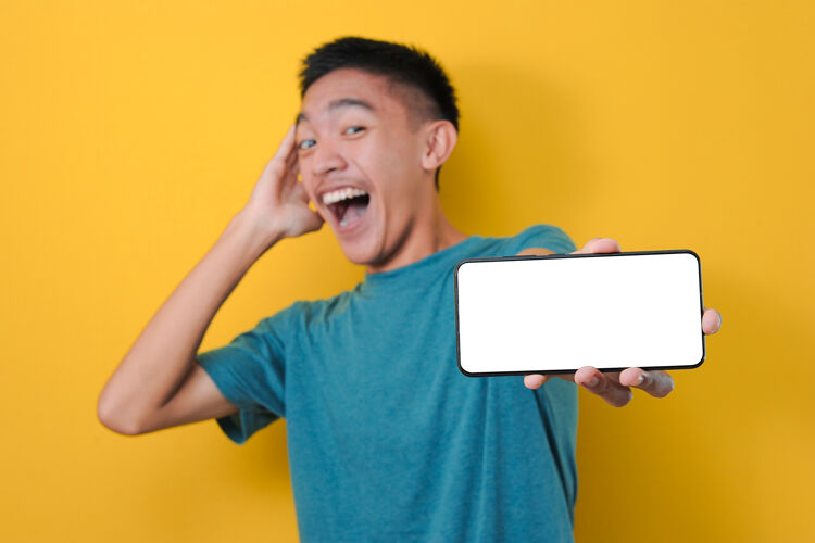 触摸屏快乐兴奋的亚洲年轻人震惊地在镜头前展示白色手机屏幕 隔离在黄色背景上显示移动技术