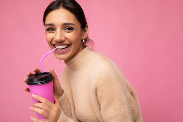 微笑迷人的年轻快乐的黑发女人穿着日常时尚的衣服隔着彩色背景墙拿着纸杯喝茶看着相机美味饮料年轻的
