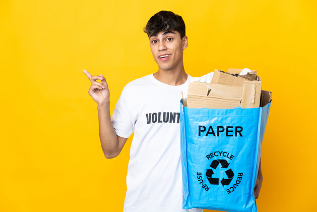 环境一个男人拿着一个装满纸的回收袋在孤立的黄色背景上进行回收 他想在举起手指的同时实现解决方案生态思考浪费