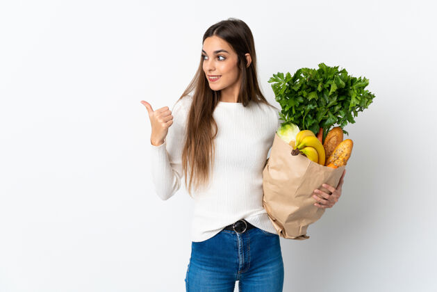 有机年轻的白种女人在买一些食物时用白色背景孤立地指着旁边的产品来呈现年轻营养表情