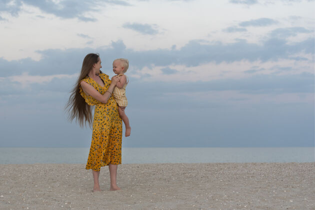自然年轻的妈妈穿着亮黄色的太阳裙 留着一头长发宝贝肖像妈妈带着孩子在海上的背景母亲童年大海