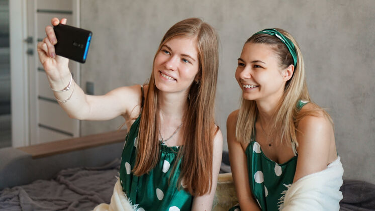生活方式两位美丽快乐的女朋友在睡衣派对上用智能手机自拍聚会自拍肖像