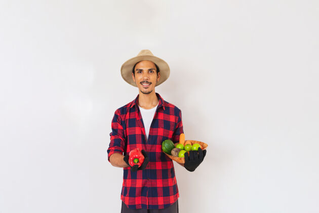 微笑一个戴着帽子和手套的黑人农民手里拿着一篮子蔬菜（胡萝卜 柠檬 西红柿 佛手瓜和甜菜） 背景是白色的携带农业西红柿