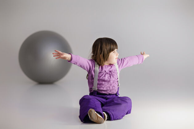 快乐小可爱的女孩 穿着紫色衣服 双臂张开 灰色背景上有一个健身用的大球颜色健康欢呼