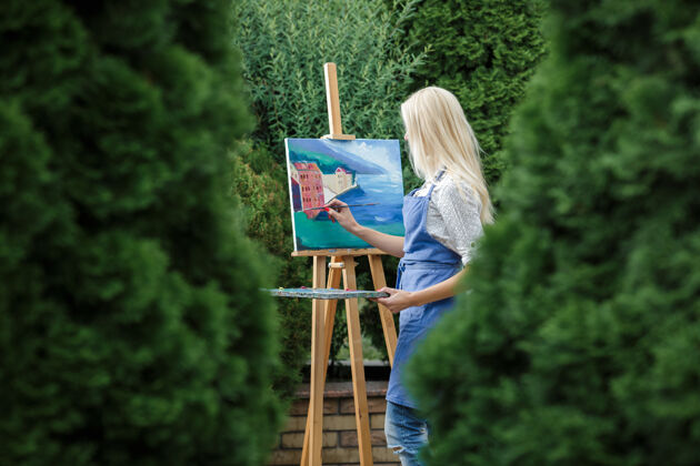 自然美丽的金发女画家手里拿着画笔在花园里的画布上画画金发女郎艺术品花园