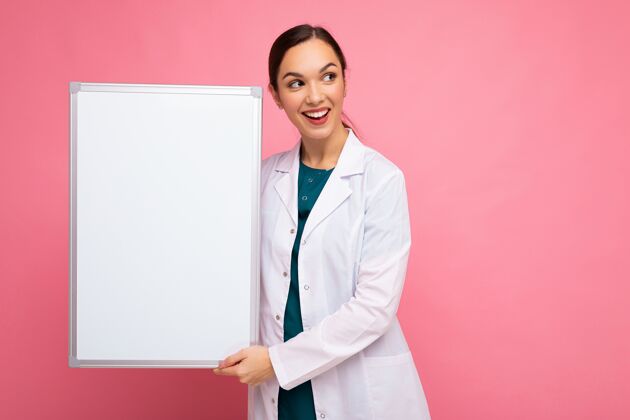 流行病有吸引力的年轻女性正面黑发 身穿白色医用外套 手持白色磁性板 在粉色背景上隔离 留有复印空间青年医生保健