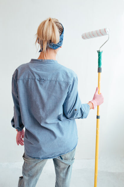 室内一个穿着蓝色衬衫和牛仔裤的女孩拿着一个粉刷墙壁的滚筒背部工具举行
