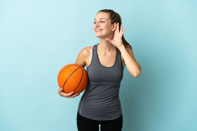 健康一个在蓝色背景上打篮球的年轻女子 手放在耳朵上听东西篮球比赛运动员