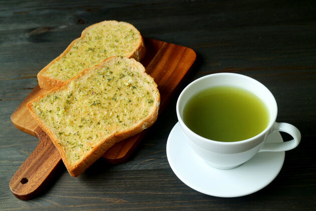 抹茶蒜黄油在面包板上烤着 红木桌上放着一杯热绿茶饮料令人垂涎三尺茶
