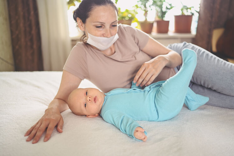 保护面罩戴着防护面罩的年轻母亲抱着一个穿着蓝色连体衣的可爱新生婴儿 抱着躺在床上的他可爱债券抱
