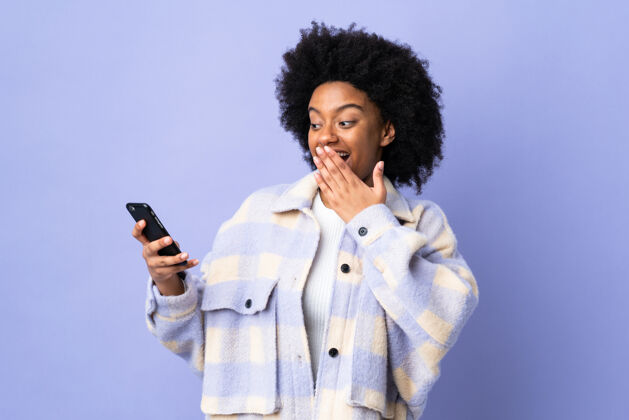 通讯年轻的非洲裔美国女子用手机孤立在紫色上 脸上带着惊讶和震惊的表情发生担心震惊