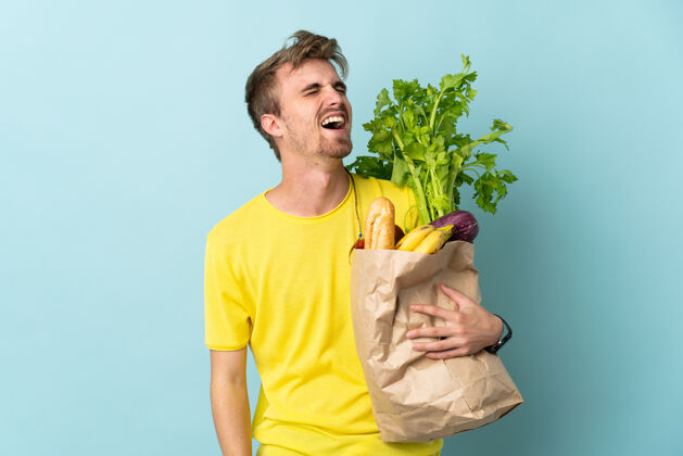 生的一个金发碧眼的人拿着一袋外卖的食物 孤立地笑着对蓝色膳食营养青年